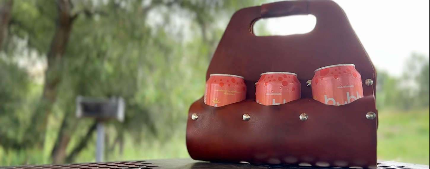 LEATHER 6 PACK HOLDER ON TABLE WITH GRILL AND TREES IN THE BACKGROUND